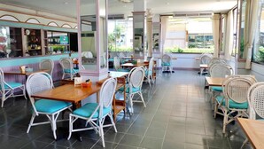 Restaurante com cadeiras de vime branco e almofadas azul-esverdeadas, piso de azulejos e amplas janelas com vista para o exterior.