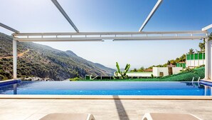 Zona de piscina con vistas despejadas a un paisaje montañoso, área de descanso techada y un edificio con detalles verdes.