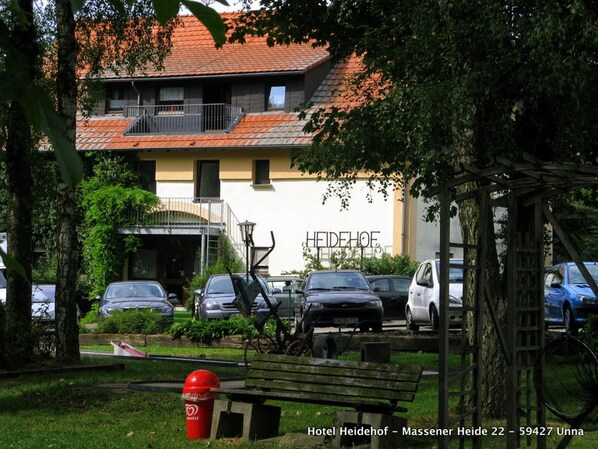 A hotel with a tiled roof, parked cars, and a red trash bin. - Hotel Heidehof (Unna)