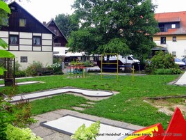 Un parque infantil con un tobogán amarillo y rojo, una pequeña estructura para escalar y un columpio verde y amarillo en un área de césped, con un edificio al fondo.