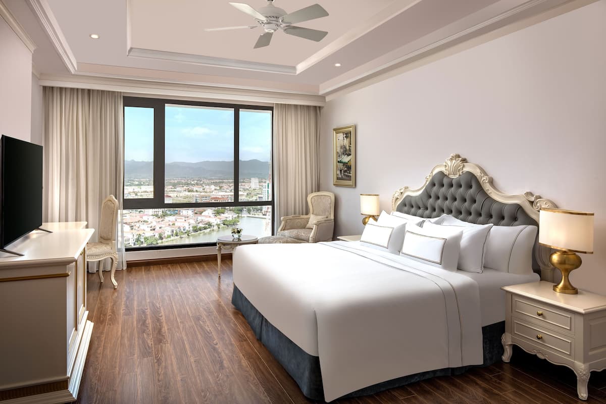 A hotel room with a large bed, a desk, a chair, a TV, and a view of a cityscape through large windows.