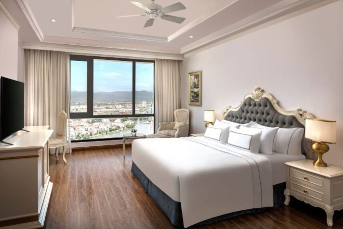 A hotel room with a large bed, a desk, a chair, a TV, and a view of a cityscape through large windows.