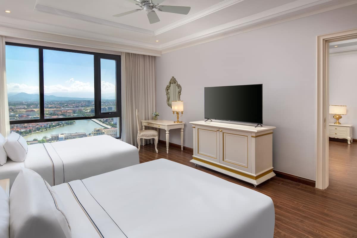 A hotel room with two beds, a desk, a TV, and a large window with a city view.