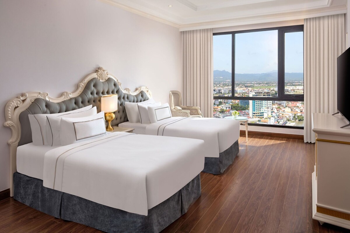 A hotel room with two beds, a large window with a city view, a TV, and a desk.