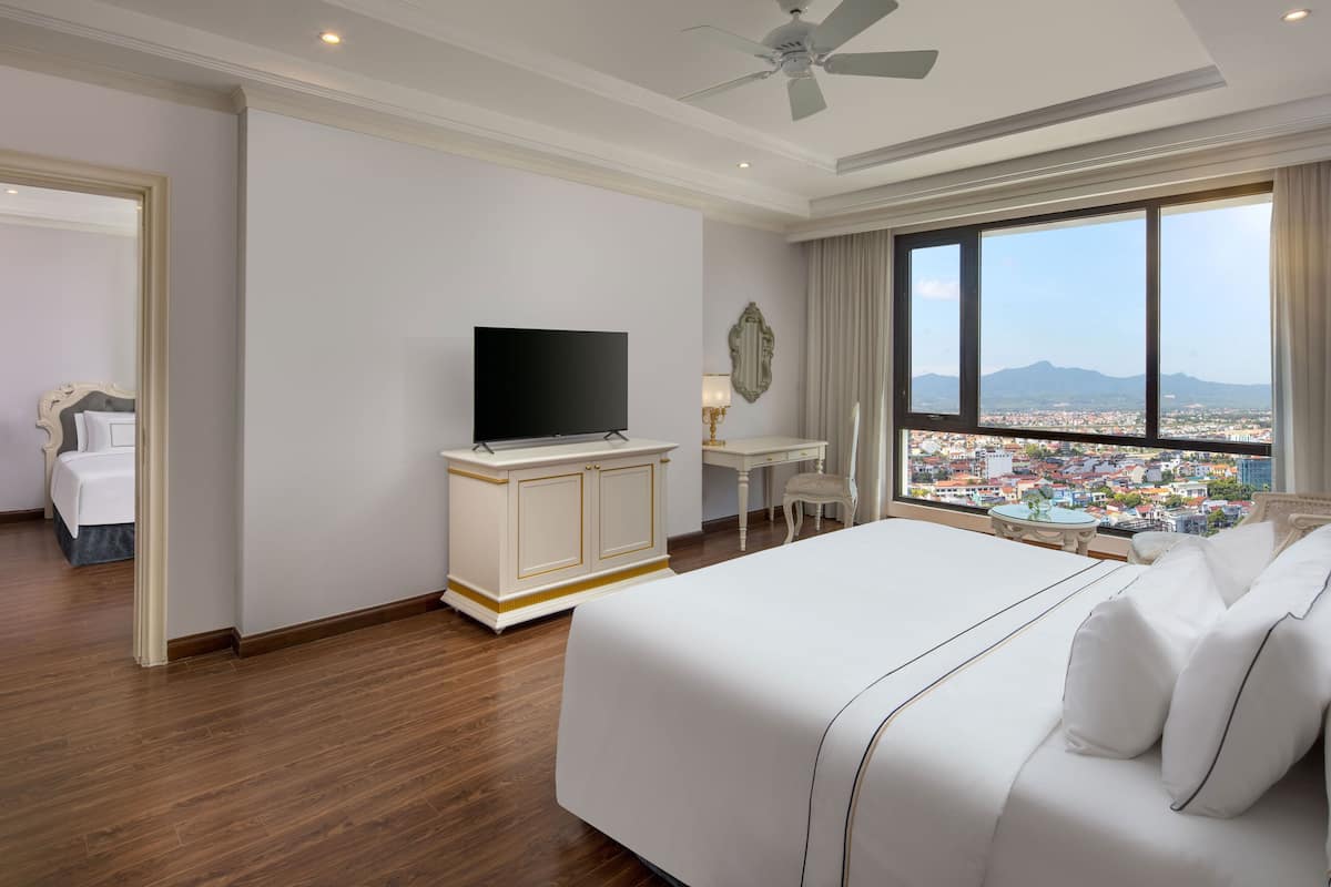 A hotel room with a large bed, a television, a desk with a chair, and a view of a cityscape through large windows.