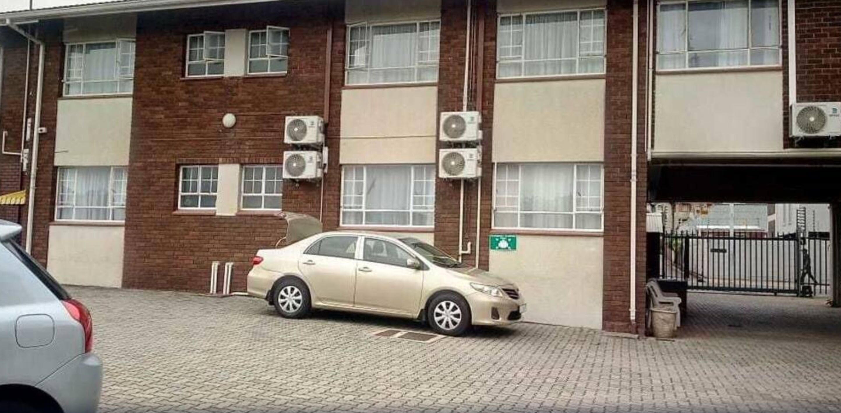 Un edificio de dos pisos con varias unidades de aire acondicionado, un coche estacionado adelante y una entrada con reja.