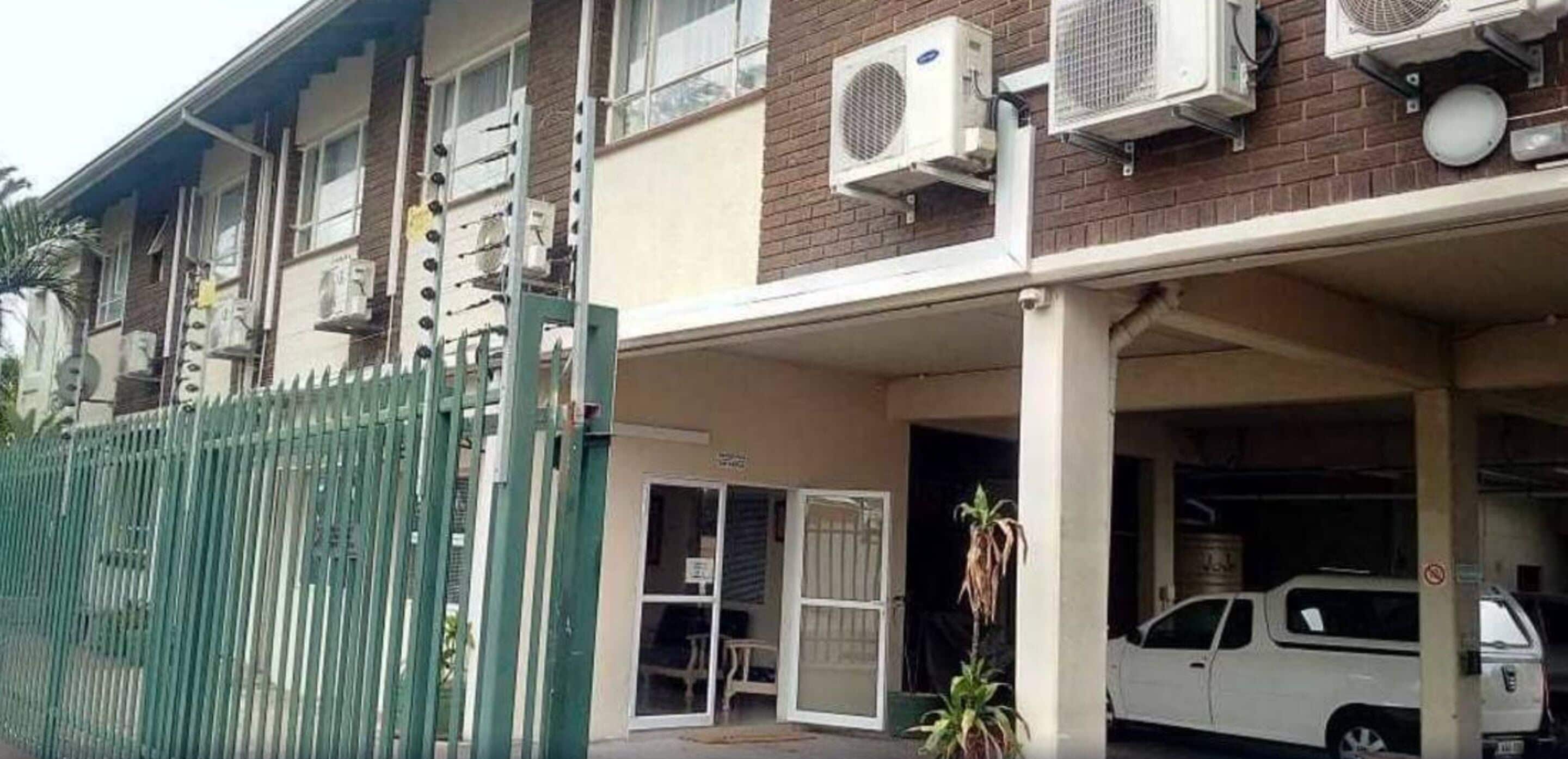 Um edifício residencial com vários aparelhos de ar condicionado, portão fechado e um SUV branco estacionado.