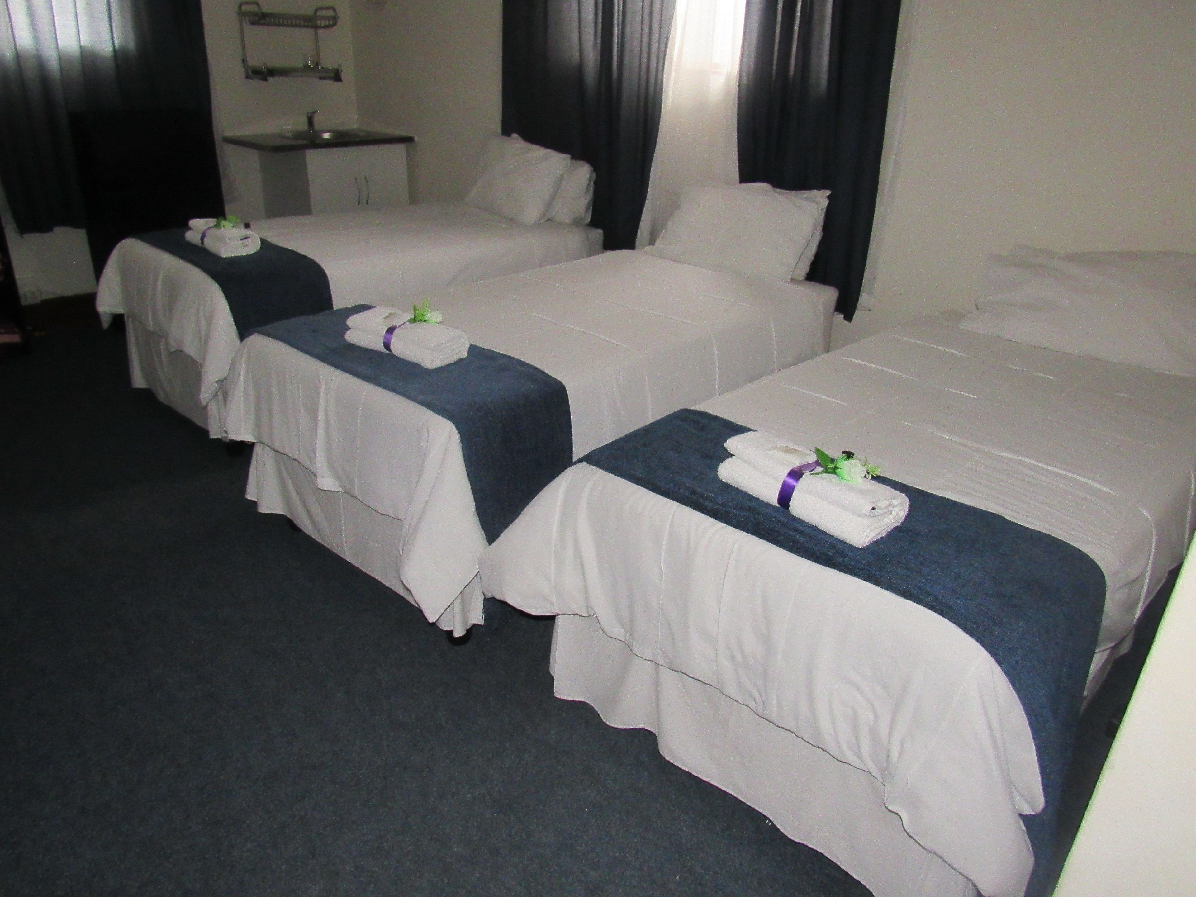 A hotel room with two single beds, each with a neatly folded towel and a small flower. There is a bedside table with a lamp and a mirror.