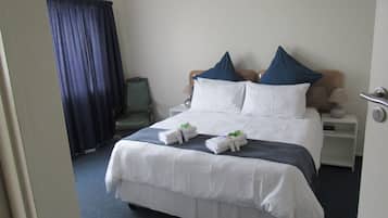 Habitación de hotel con cama, dos almohadas, una silla, mesita de noche con lámpara y ventana con cortinas.