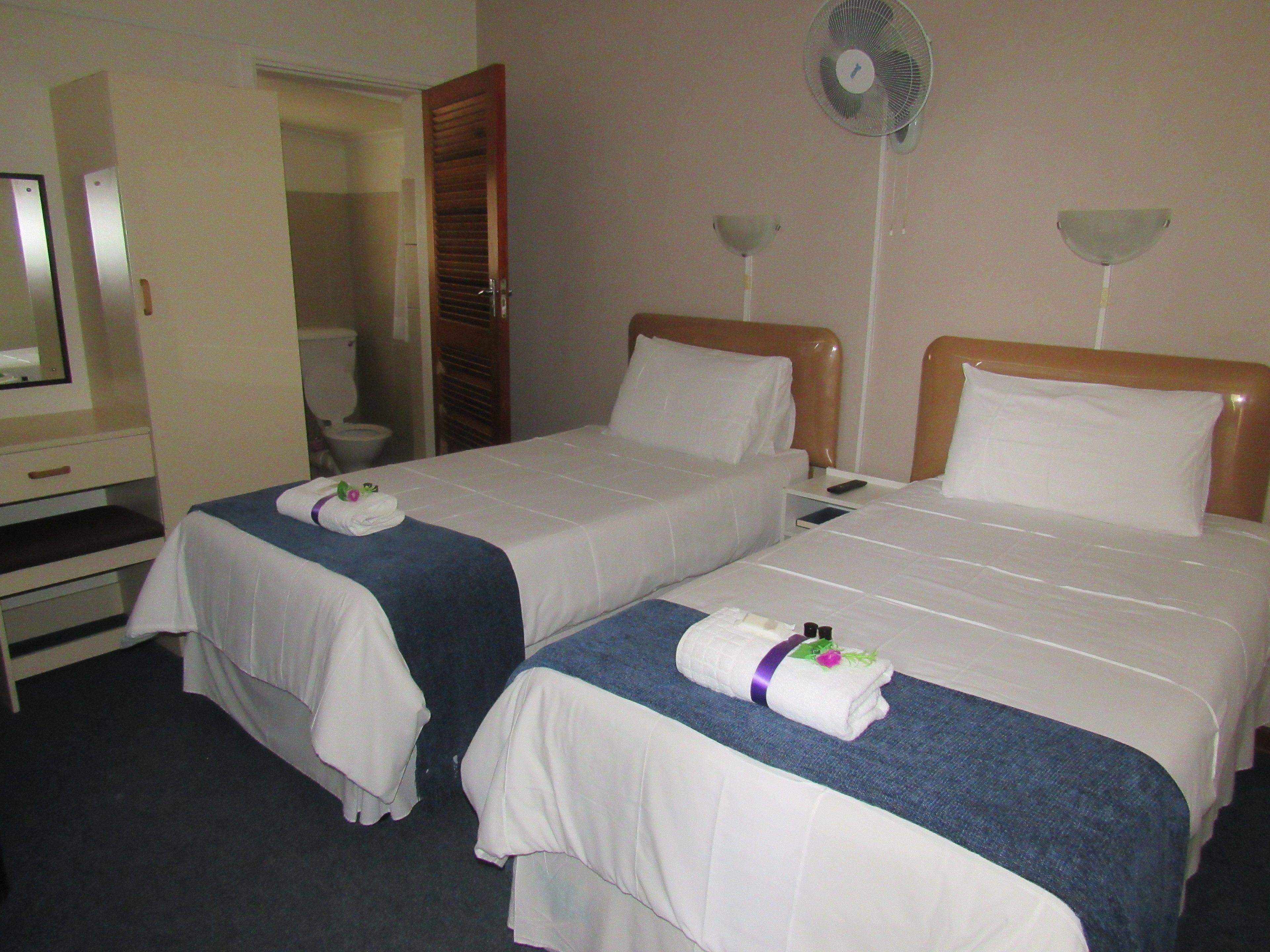 A hotel room with two beds, a desk, a mirror, a fan, and a toilet.