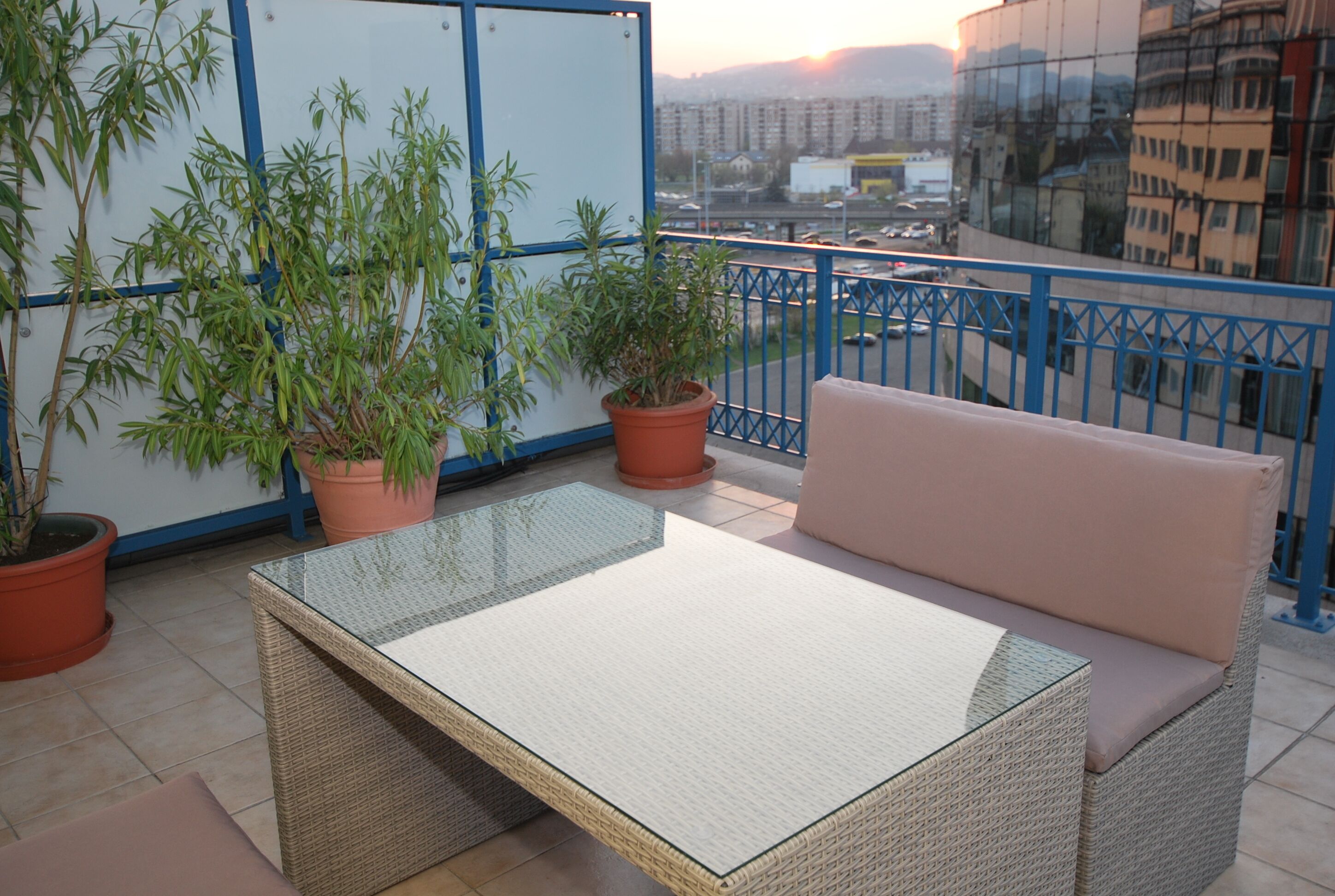 Un balcón con una mesa de vidrio, sofá de mimbre y plantas en macetas.