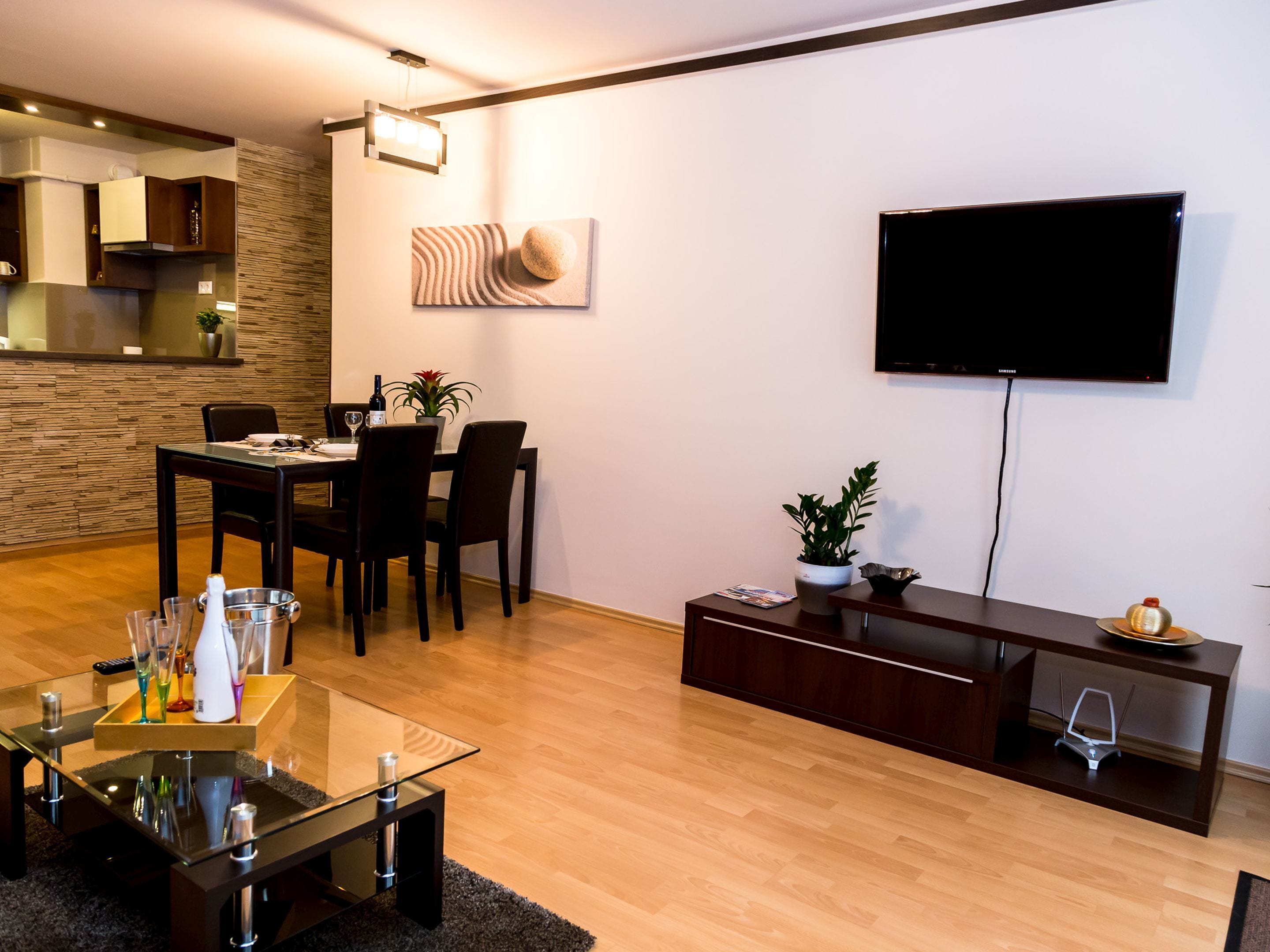 A modern living room with a dining area, a flat-screen TV mounted on the wall, and a wooden floor.