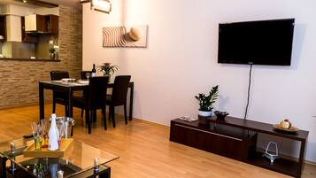 A modern living room with a dining area, a flat-screen TV mounted on the wall, and a wooden floor.