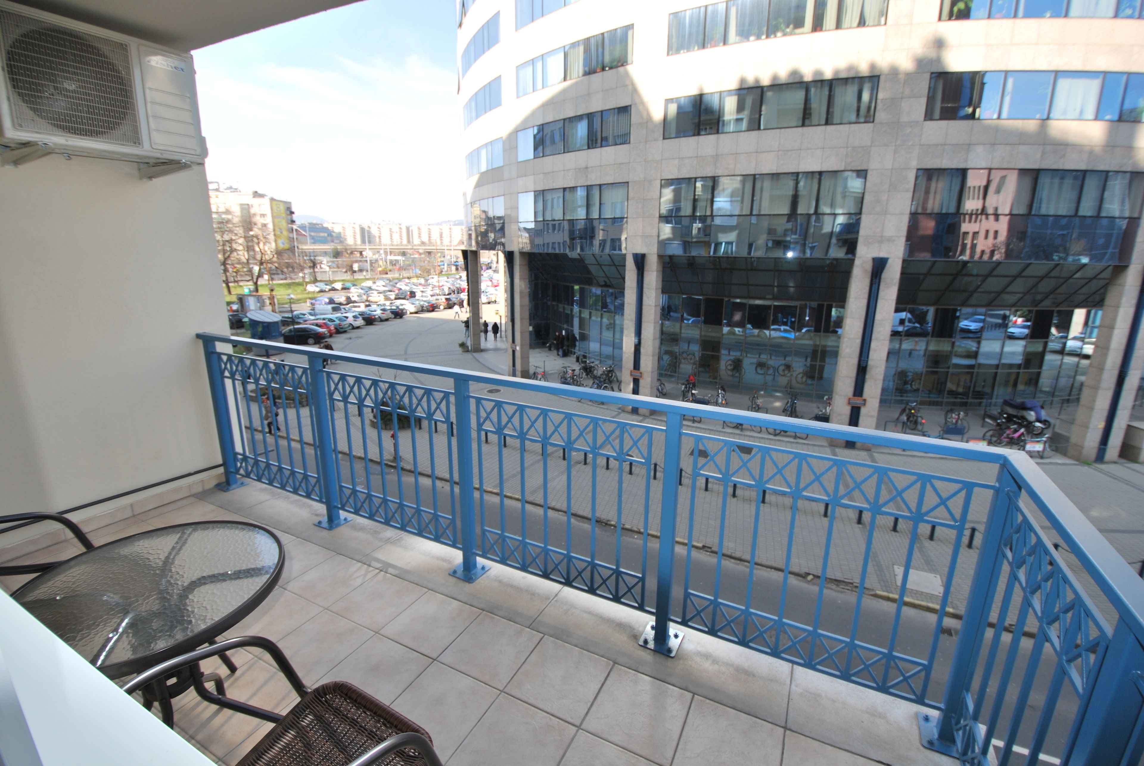 A balcony with a blue railing overlooking a parking lot and a modern building with large windows.