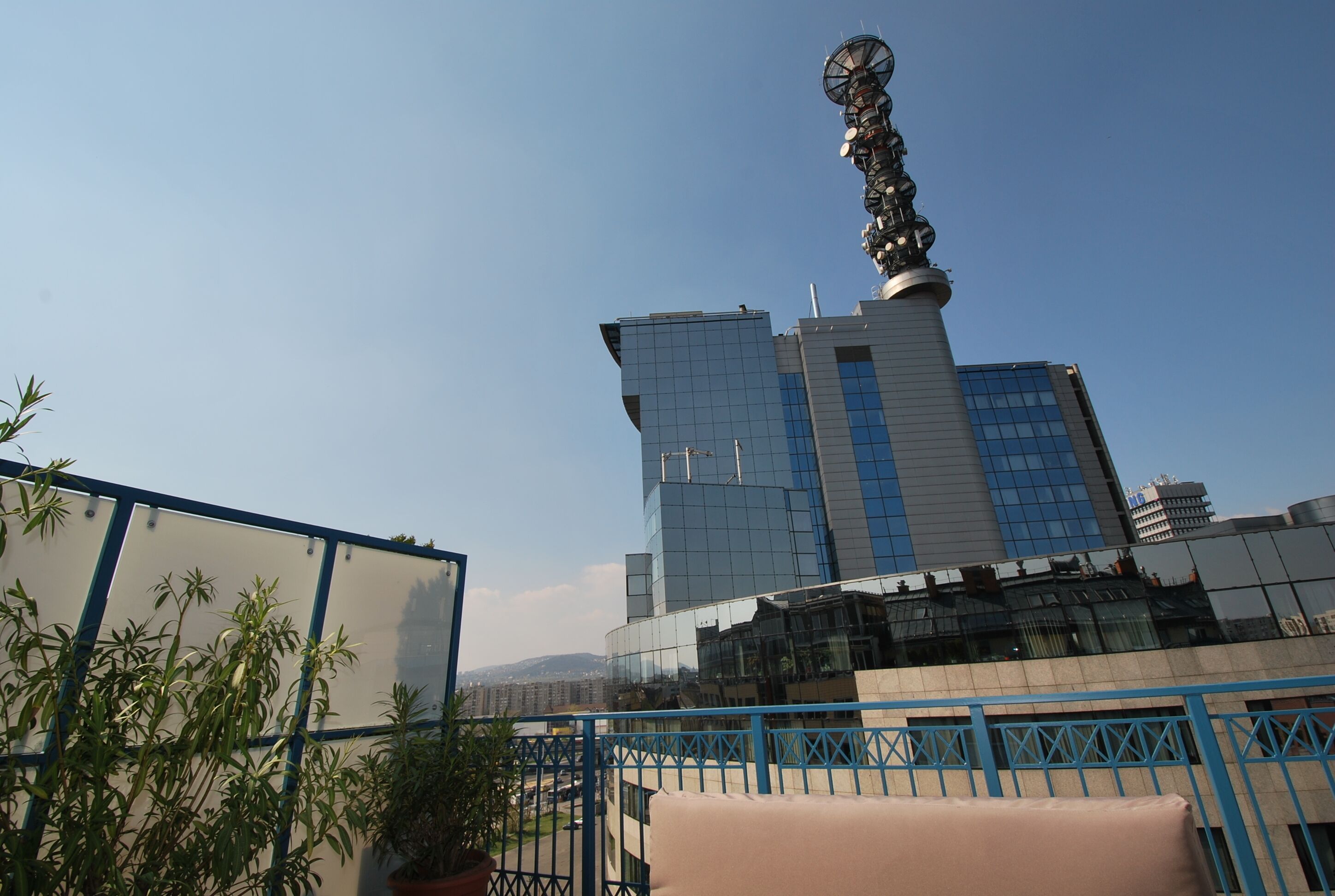 Un edificio moderno con una estructura de torre distintiva, rodeado de plantas en macetas y una barandilla azul.