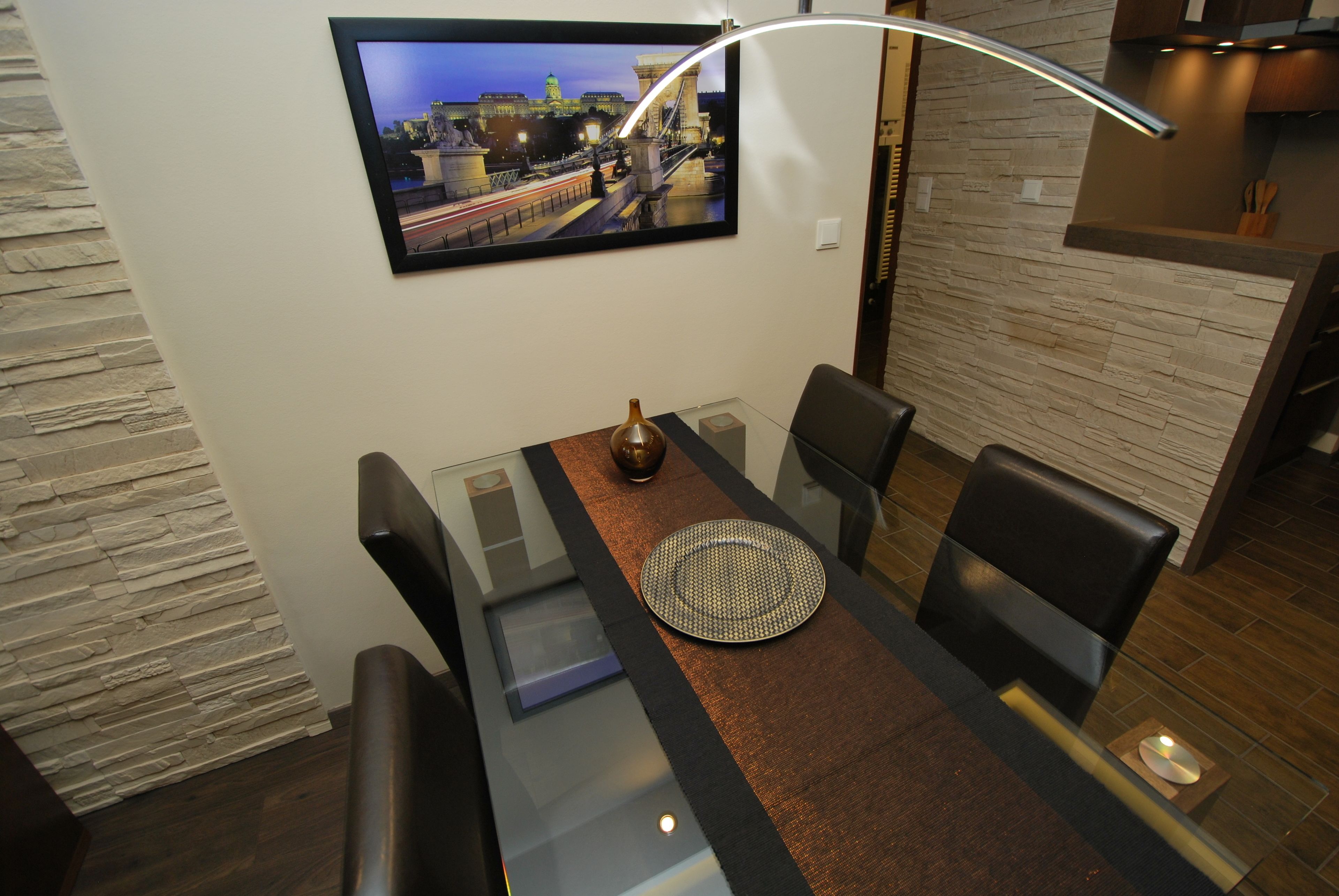 Een moderne eetruimte met een glazen tafel, zwarte stoelen, een geweven placemat en een stadsgezicht schilderij aan de muur.