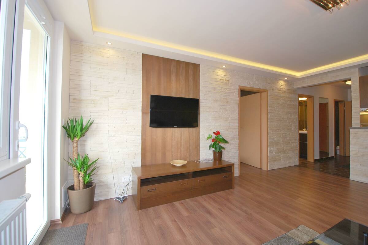 A modern living room with a flat-screen TV mounted on a stone wall, wooden flooring, and a built-in entertainment unit with a potted plant.