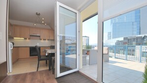 Une cuisine moderne avec des meubles en bois, un coin repas avec une table et des chaises, et un balcon offrant une vue sur un immeuble et une rambarde bleue.