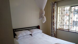 A neatly made bed with white linens and a folded towel, a window with metal bars, and a curtain with a geometric pattern. - Kadana Bed & Breakfast - Adults Only (Nairobi)