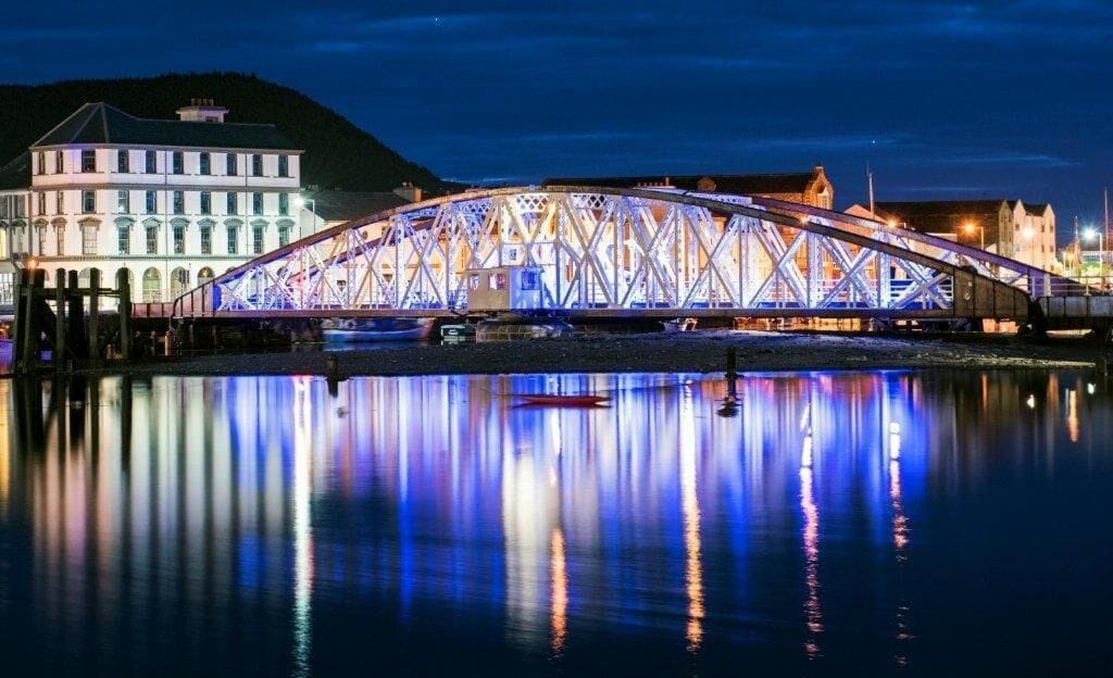 A large, illuminated bridge with a symmetrical truss design, spanning over a body of water with a calm reflection.