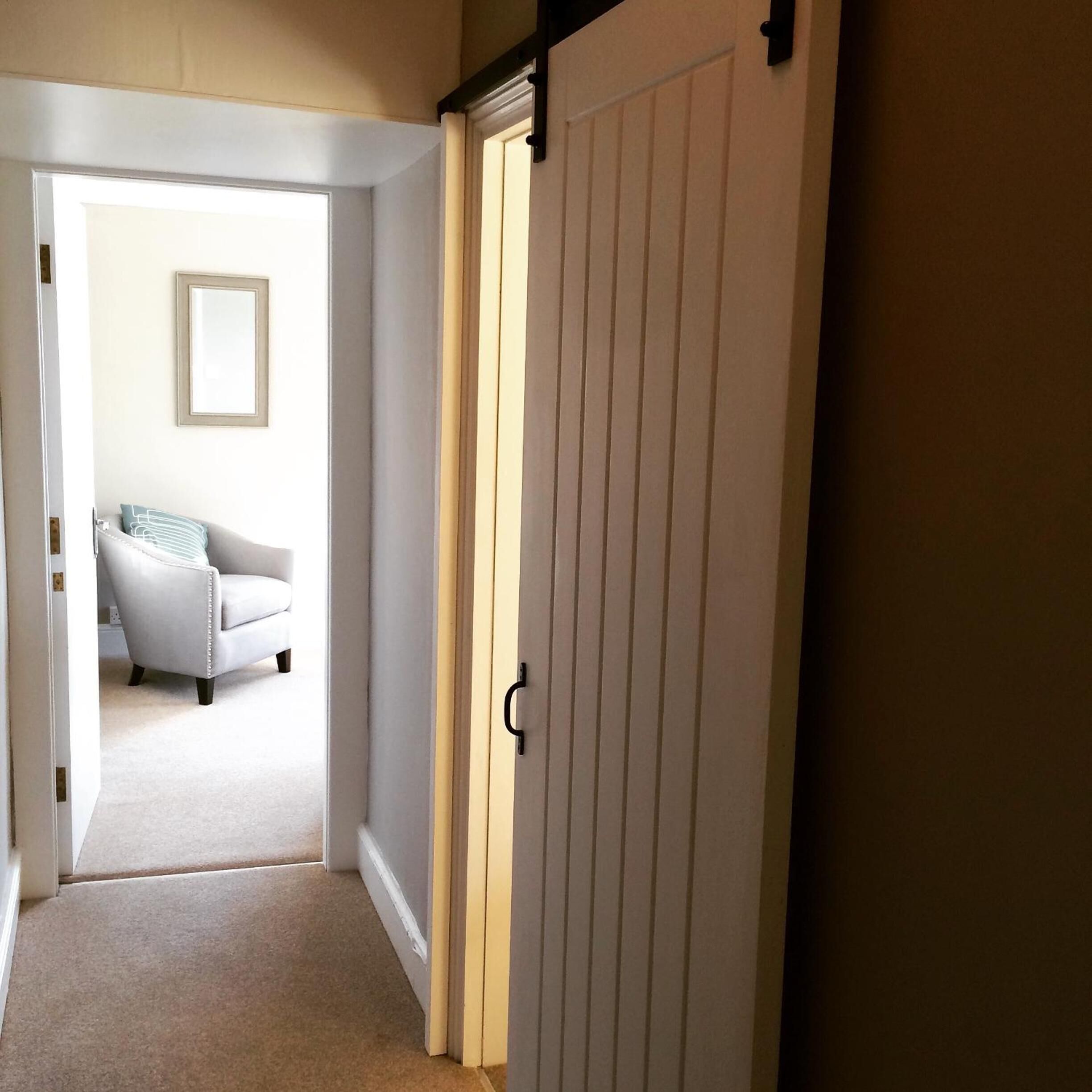 Un couloir mène à une chambre dont la porte est blanche, avec un tableau encadré accroché au mur et une chaise blanche à l’intérieur.