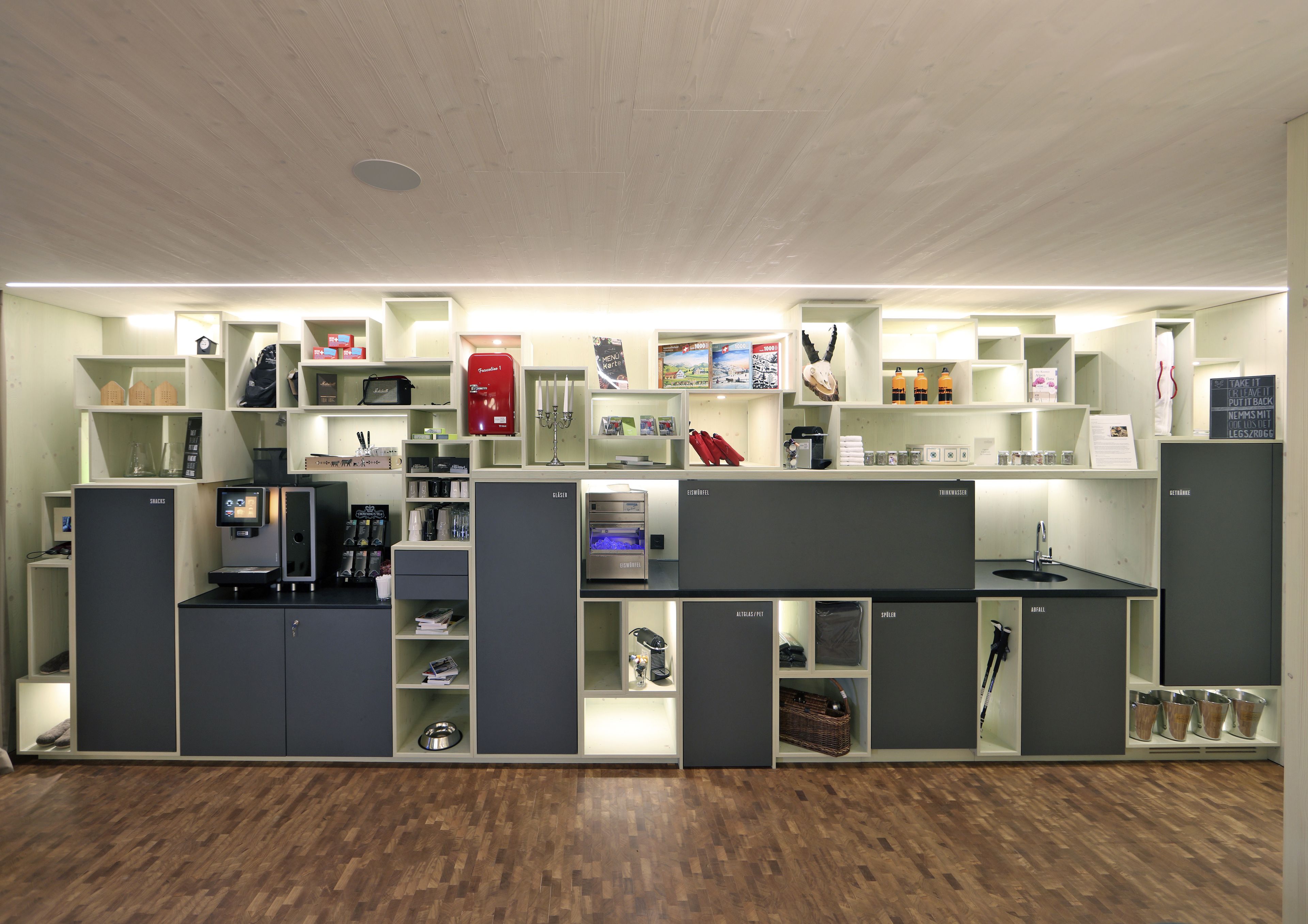 Eine moderne Hotelhalle mit Holzboden, integrierten Sitzbereichen und verschiedenen Annehmlichkeiten, darunter eine Kaffeemaschine, ein Wasserspender und ein Verkaufsautomat.