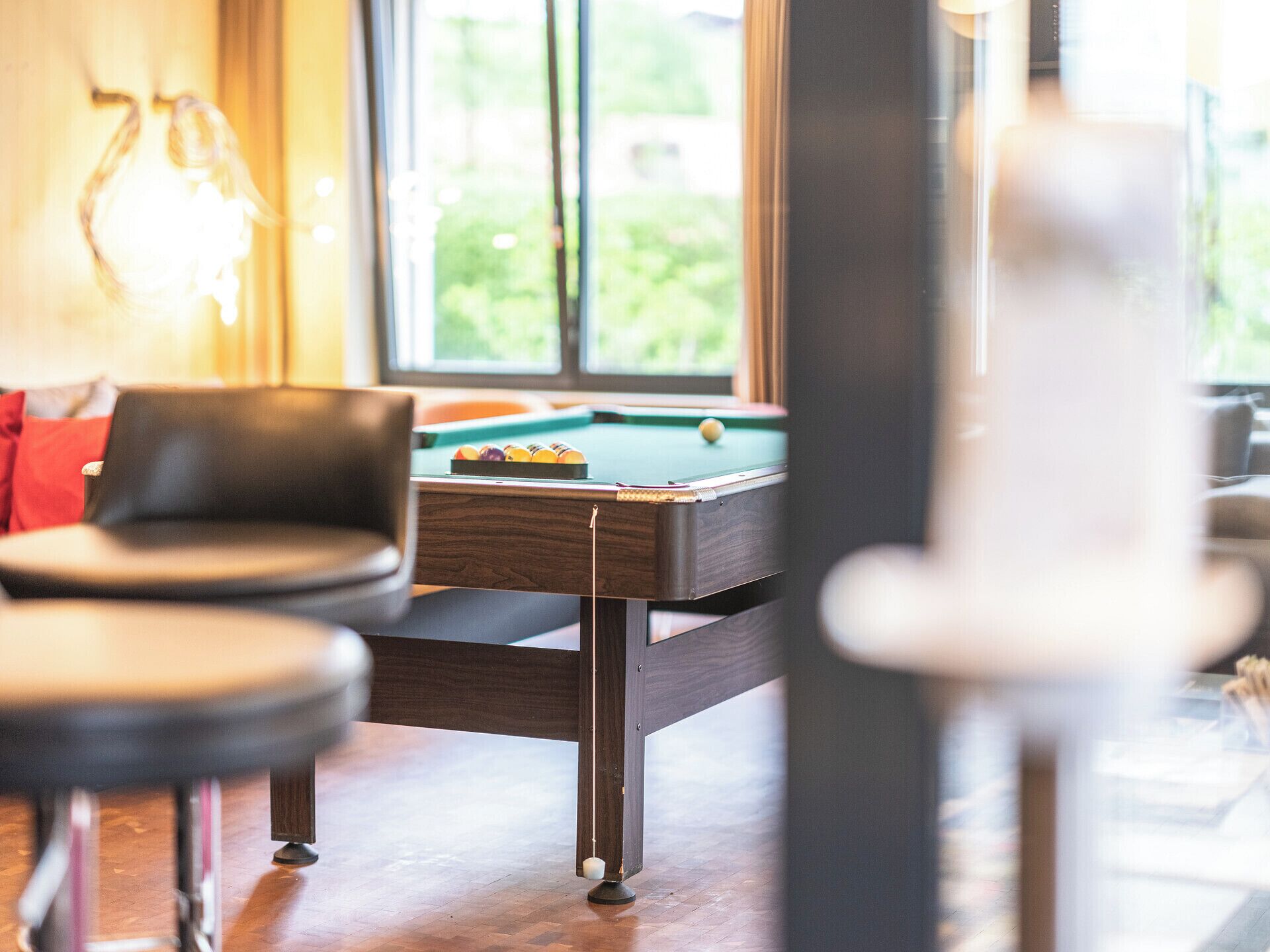 A pool table in a room with a view of greenery outside. There are two chairs and a lamp in the foreground.
