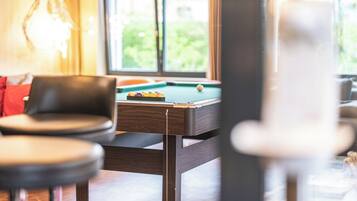 A pool table in a room with a view of greenery outside. There are two chairs and a lamp in the foreground.