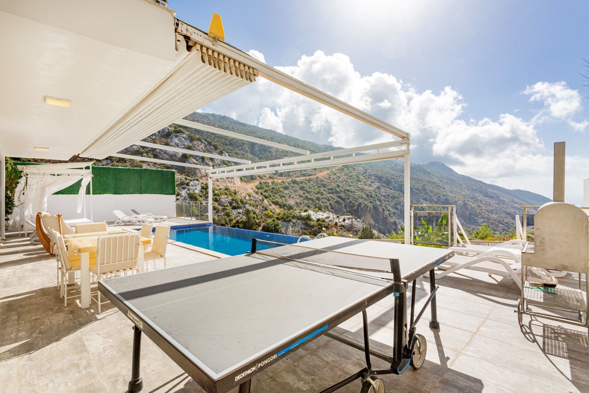 Taula de bitlles en una zona exterior il·luminada pel sol, amb vista a una muntanya, una zona de seients coberta i una piscina.