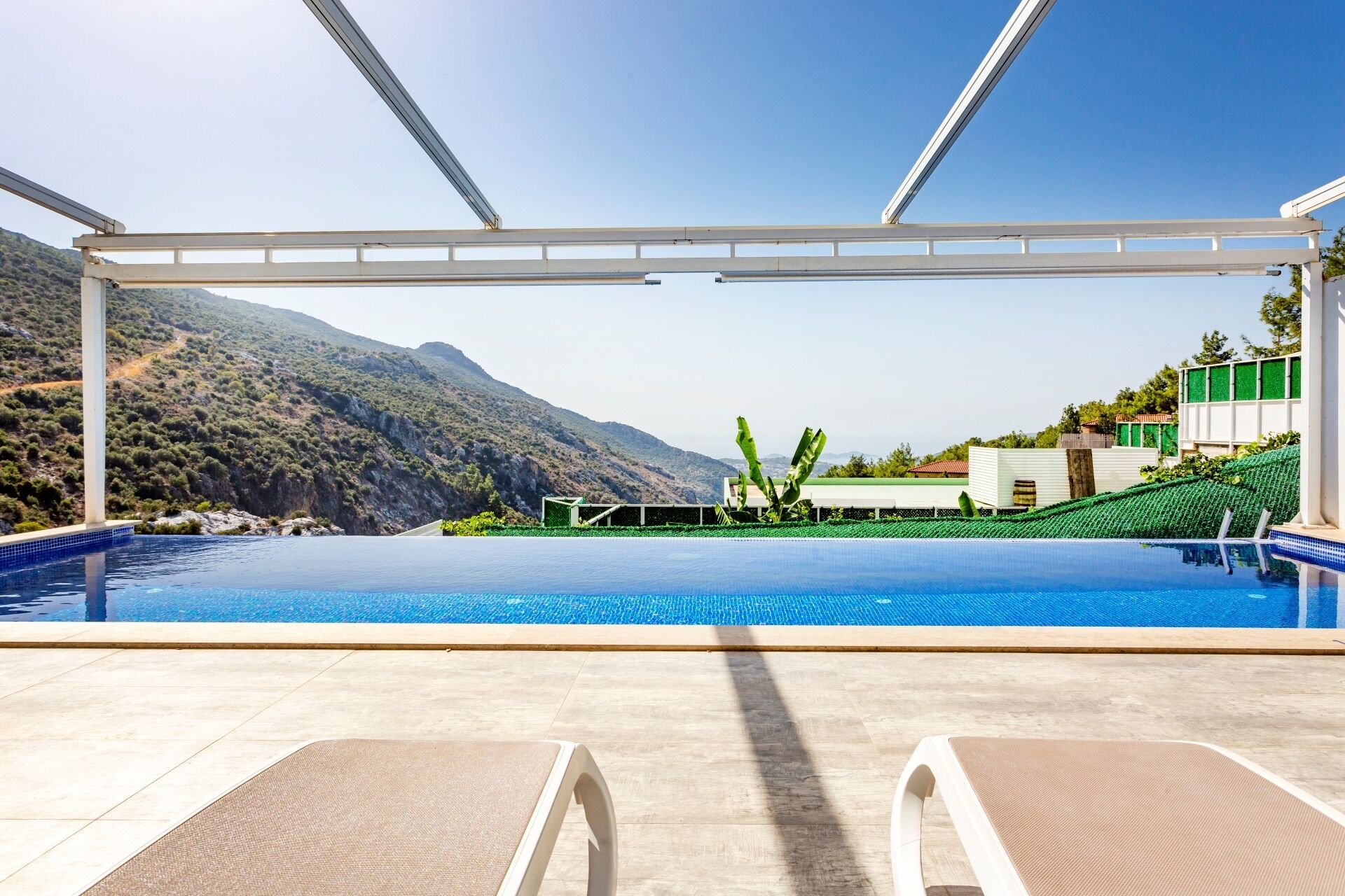 A pool area with a clear view of a mountainous landscape, a covered seating area, and a building with green accents.