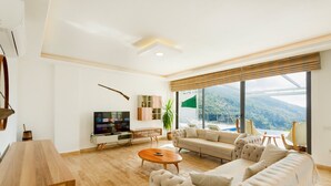 Un salon moderne avec un grand canapé, une table basse et un téléviseur. De grandes fenêtres offrent une vue sur un paysage montagneux.