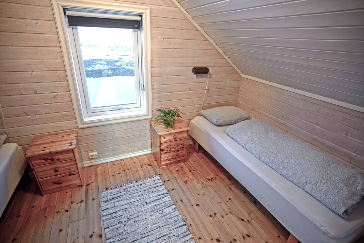 A small, cozy room with wooden walls and a sloped ceiling. It features a single bed, a small wooden nightstand with drawers, a window with a view of the outdoors, and a small potted plant on the nightstand.