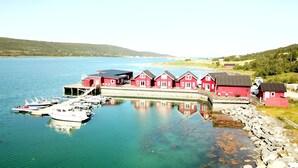 Une propriété située au bord de l’eau, avec une rangée de bâtiments rouges, un quai où sont amarrés des bateaux et une vue dégagée sur l’eau et les collines lointaines.