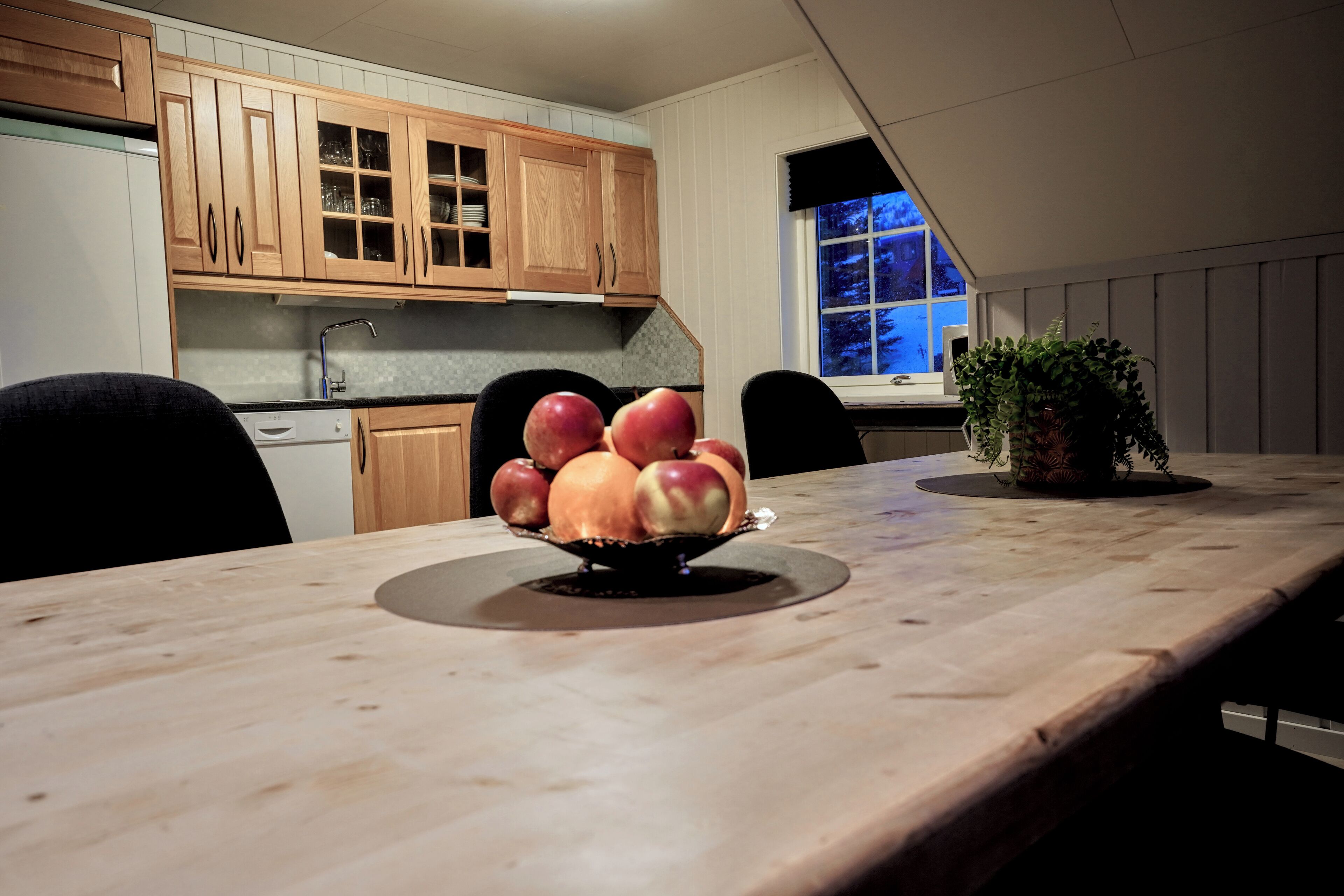 Een keuken met houten kastjes, een eettafel met een schaal fruit en een raam met uitzicht op bomen.