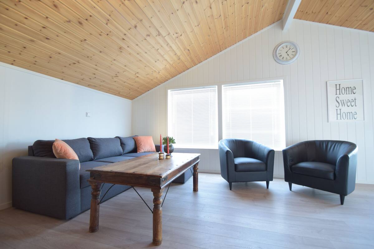A modern living room with a wooden ceiling, a sofa, two armchairs, a coffee table, and a wall clock.