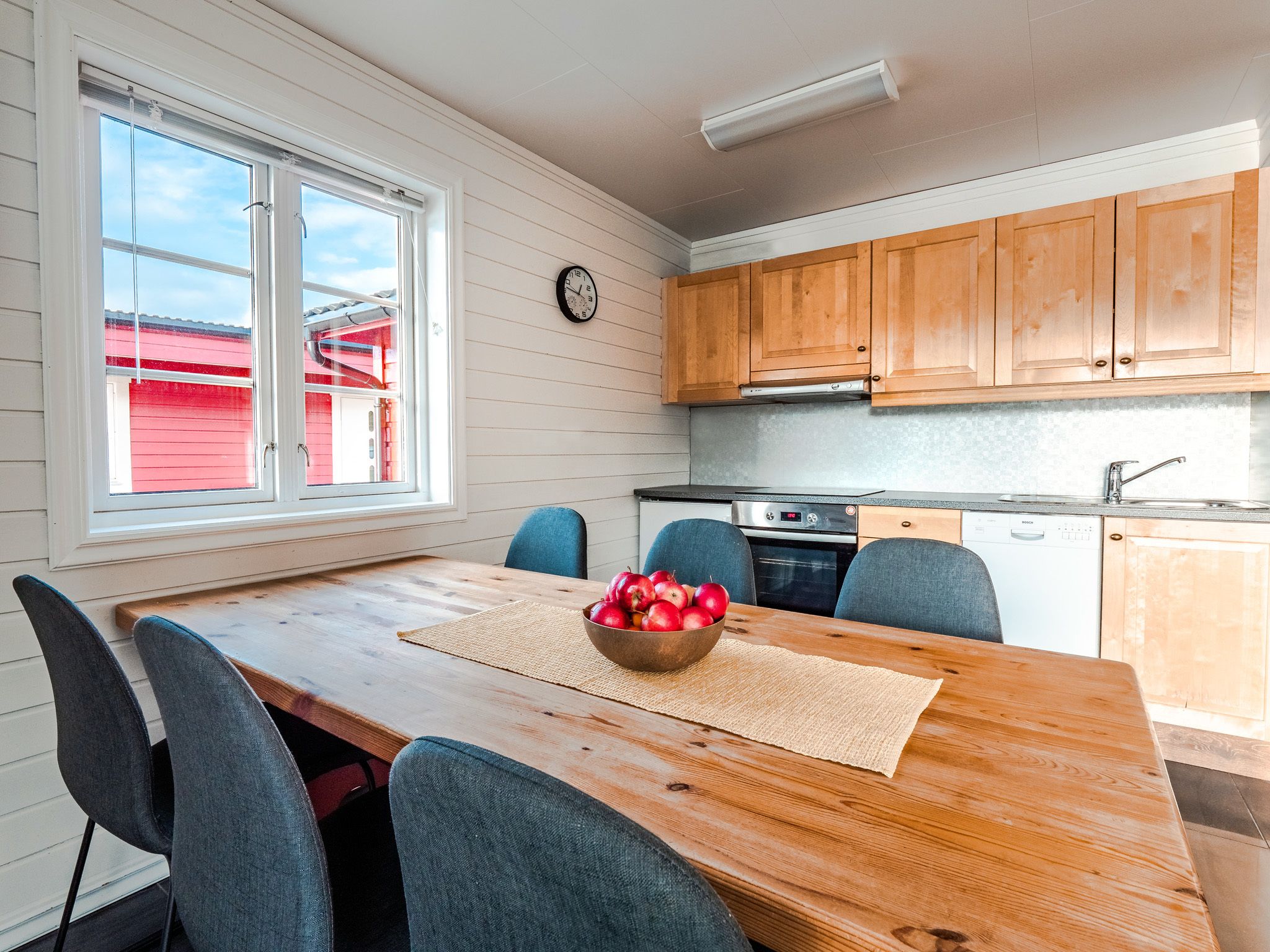 A kitchen with wooden cabinets, a dining table with chairs, a clock on the wall, and a window with a view of a red building.