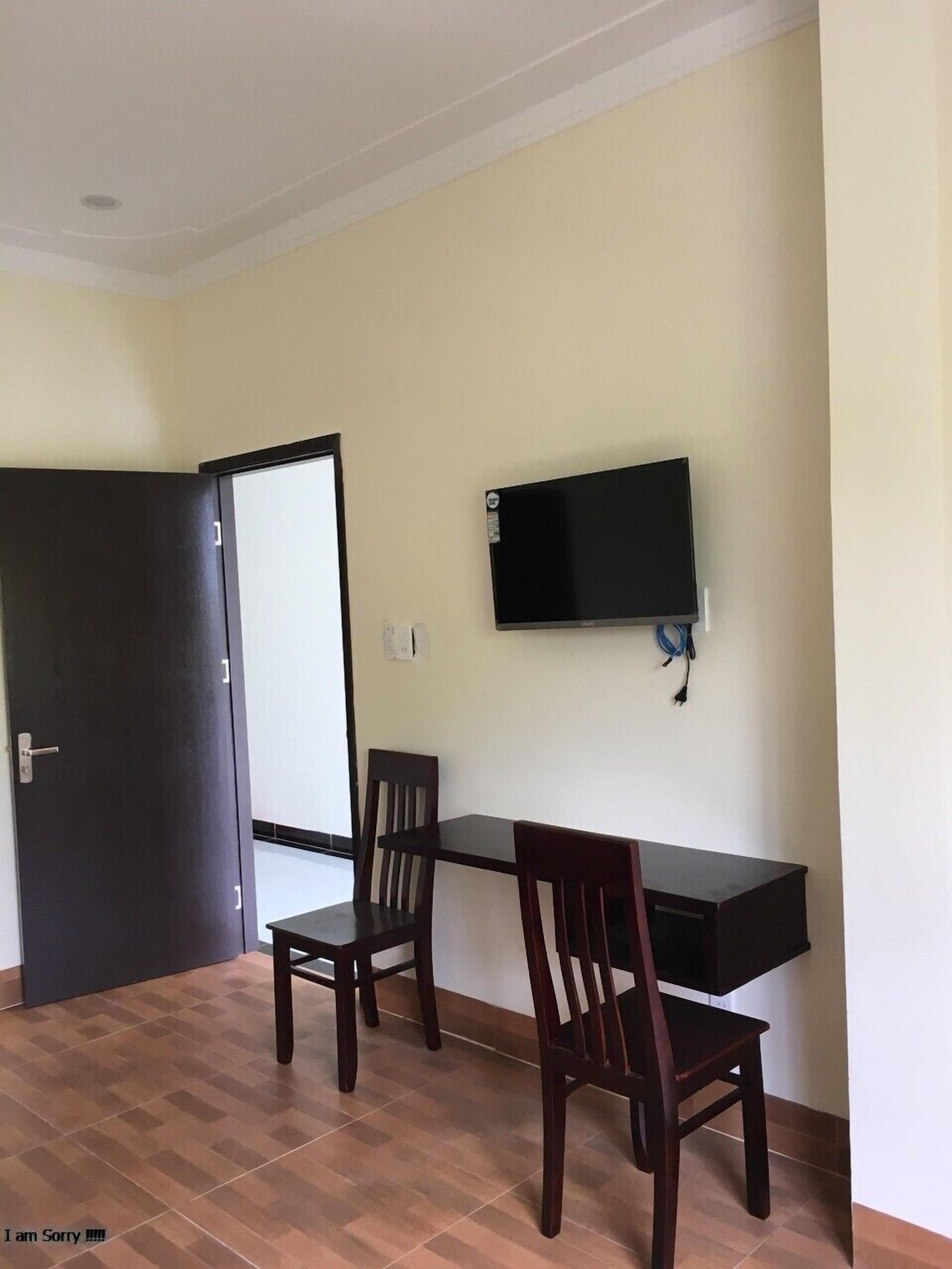 A room with a dark wooden desk, two wooden chairs, a flat-screen TV mounted on the wall, and a door with a white handle.