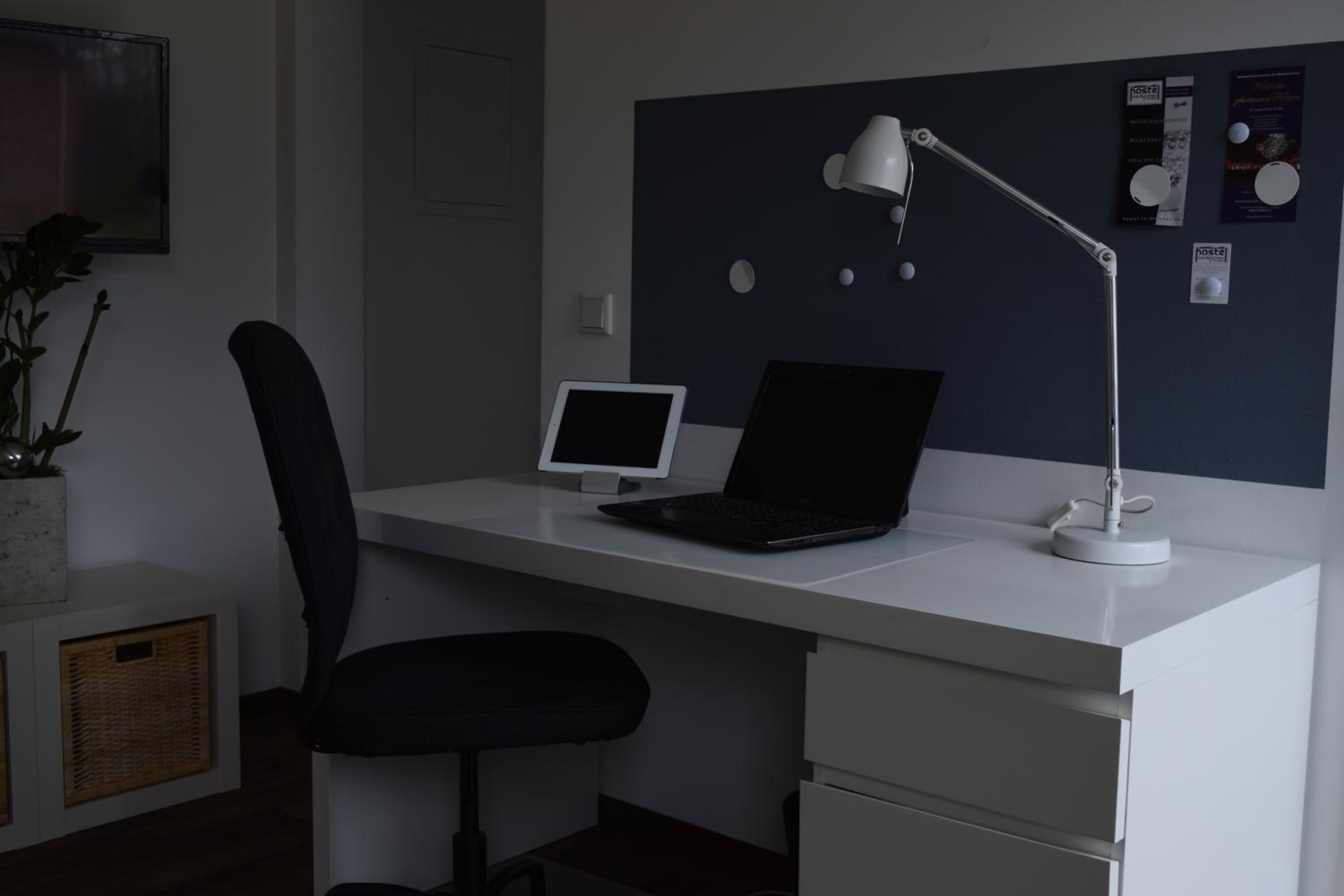A modern office desk with a laptop, a desktop monitor, and a white desk lamp. There is a black office chair and a potted plant on the left.