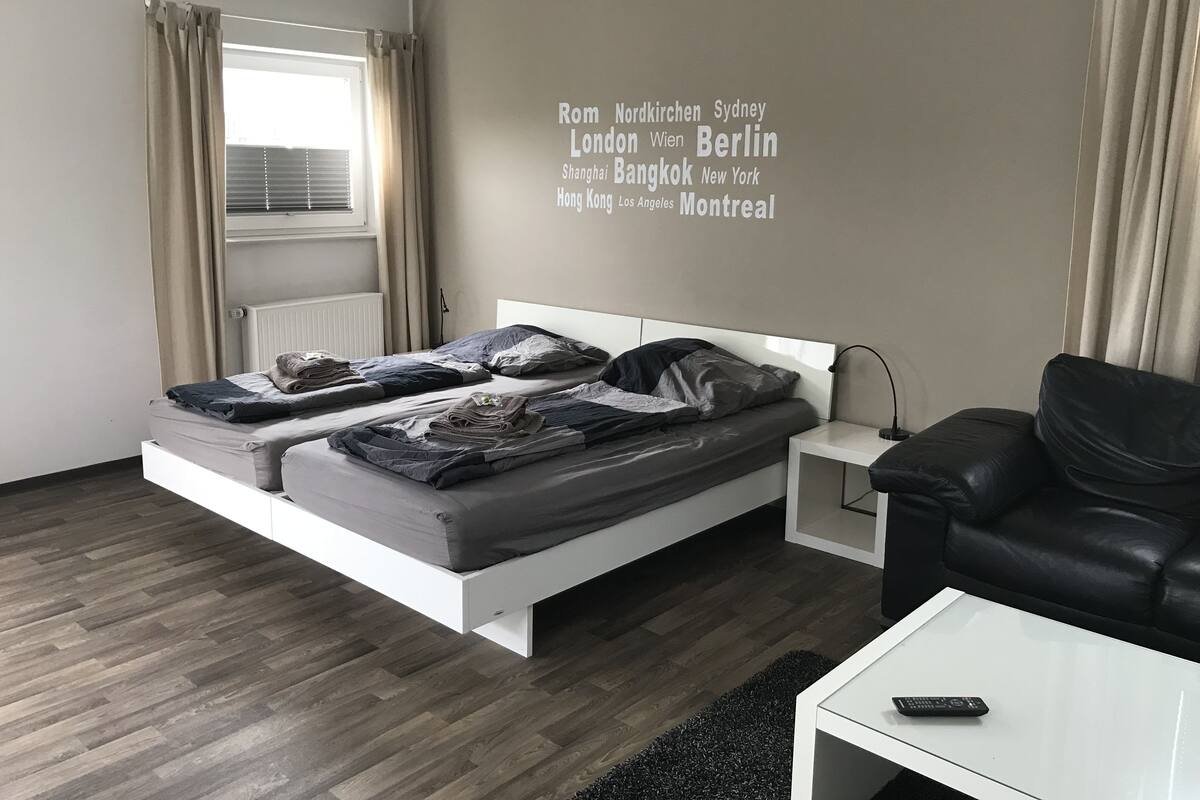 A modern hotel room with a bed, a black leather sofa, a small white table, and a wall with city names.