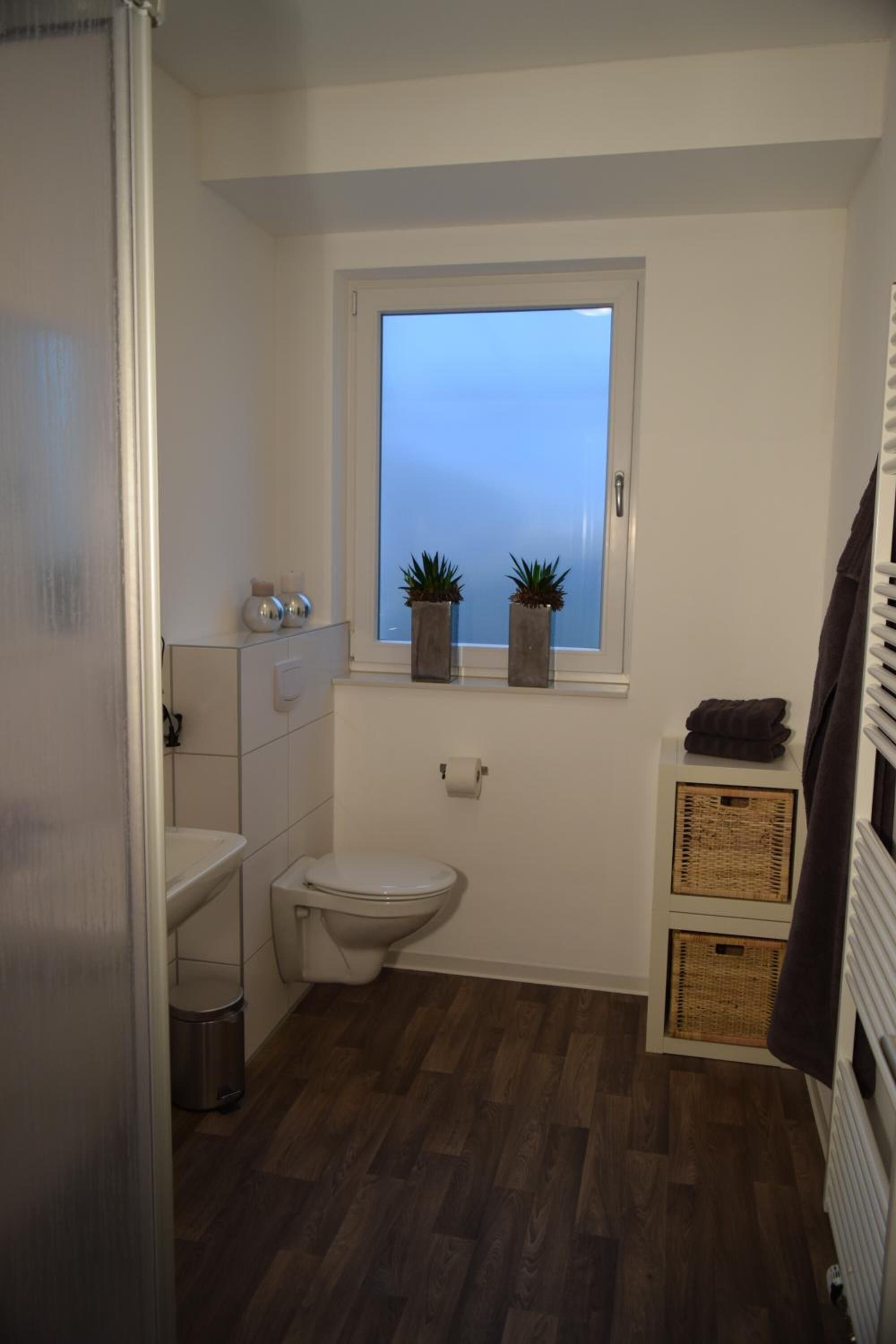 A compact bathroom with a toilet, a window, and a small shelf with plants.