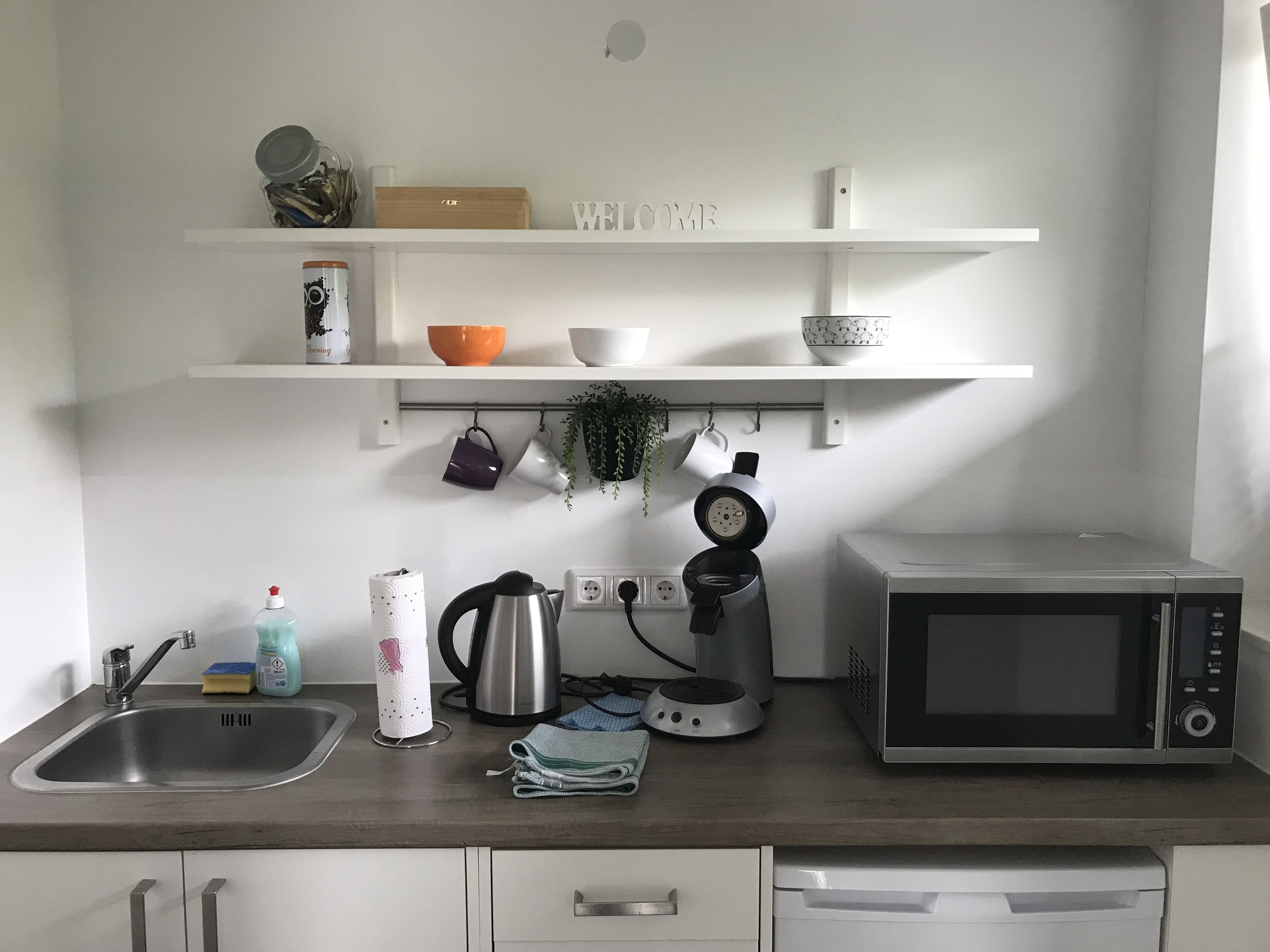 Cocina con lavamanos, hervidor, cafetera y microondas. Hay estantes sobre la encimera que contienen varios utensilios de cocina.