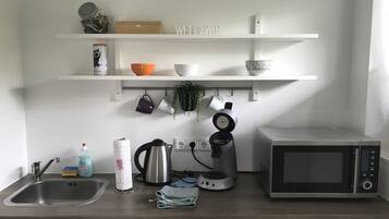 Cocina con lavamanos, hervidor, cafetera y microondas. Hay estantes sobre la encimera que contienen varios utensilios de cocina.