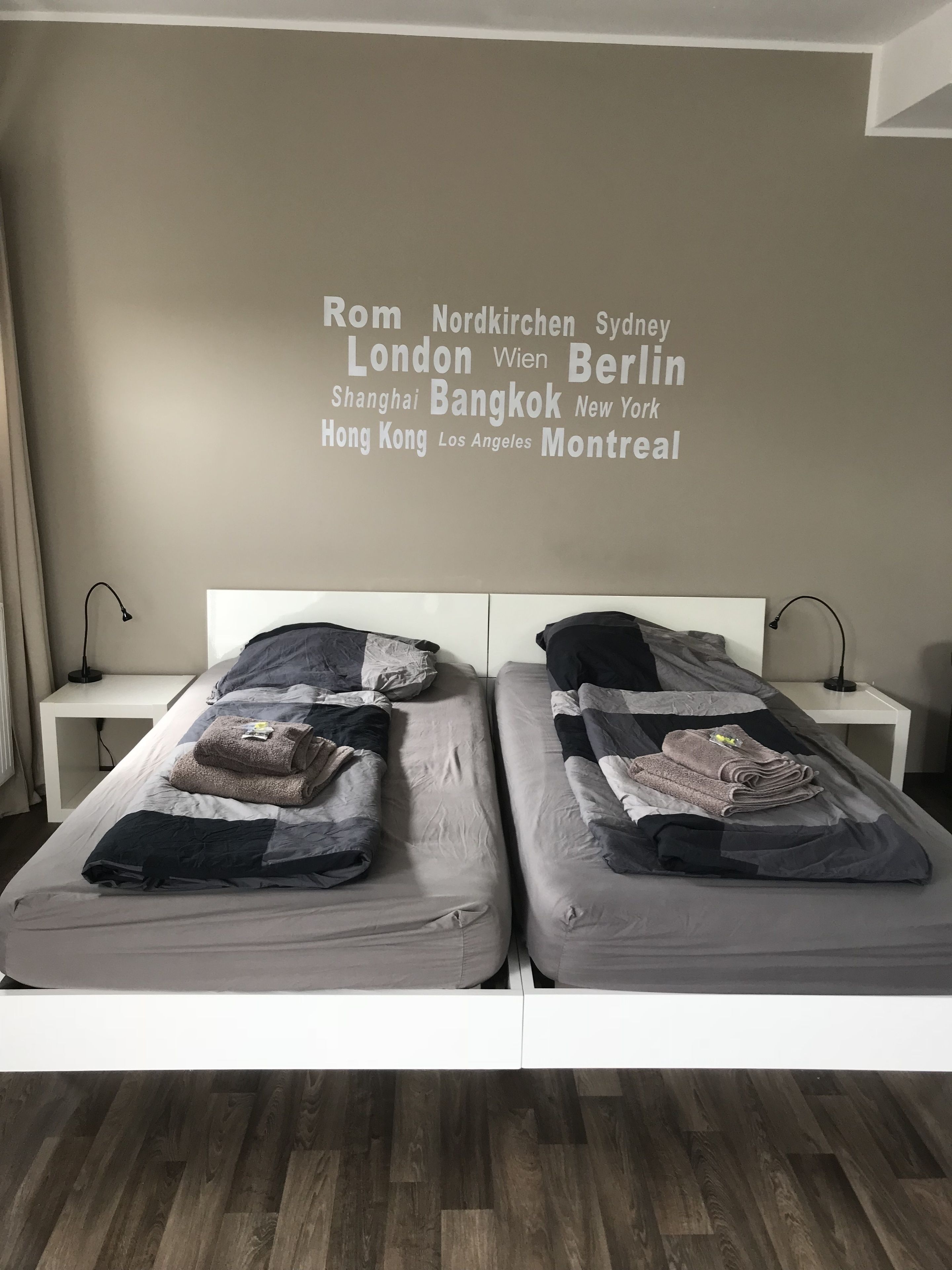 Una cama doble con edredón gris y negro, dos mesitas de noche y una pared con nombres de ciudades.