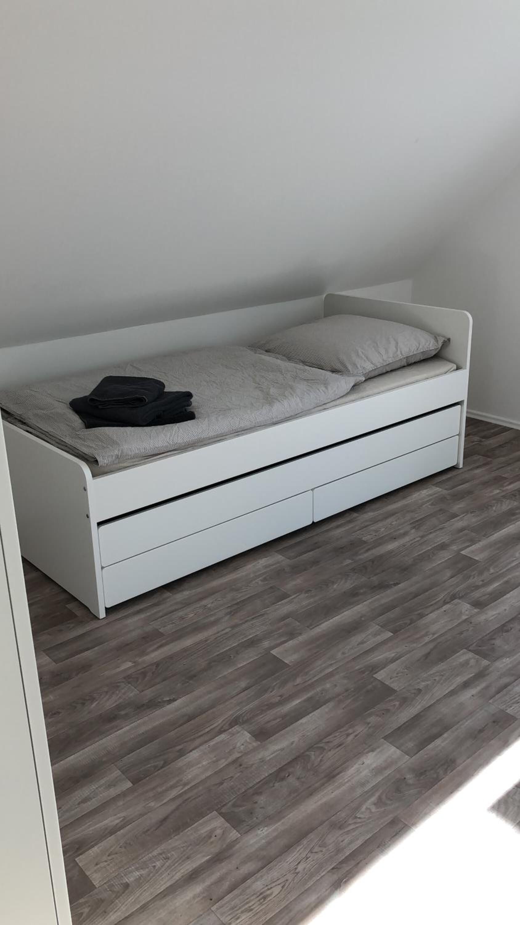 A white bed with a grey mattress and a pillow, a folded black jacket on top, and a grey blanket.