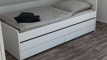 A white bed with a grey mattress and a pillow, a folded black jacket on top, and a grey blanket.