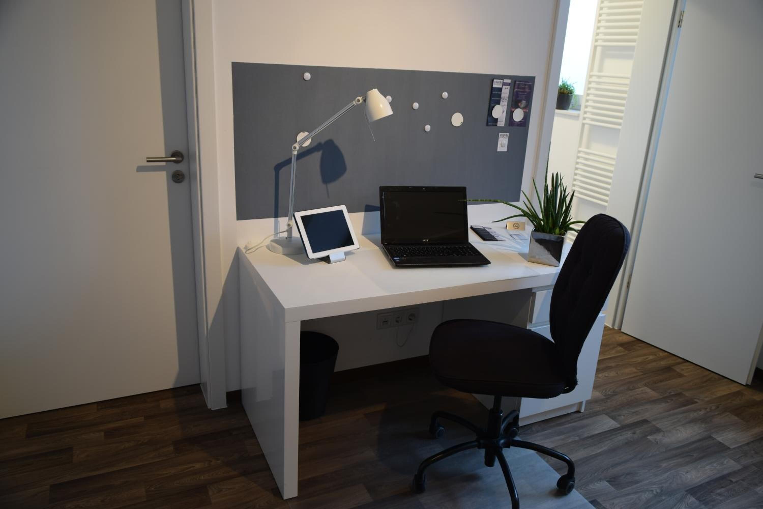 Un espacio de oficina moderno con un escritorio blanco, una computadora portátil, una tableta, una lámpara de escritorio, una silla de oficina negra y una planta en maceta.