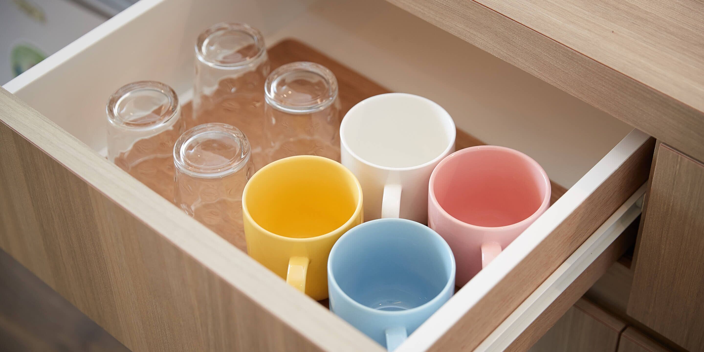 A drawer with four mugs in yellow, blue, pink, and white, and six empty glass bottles.