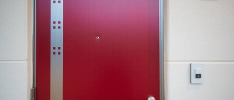 A red door with a handle, a peephole, and a light switch on the wall.