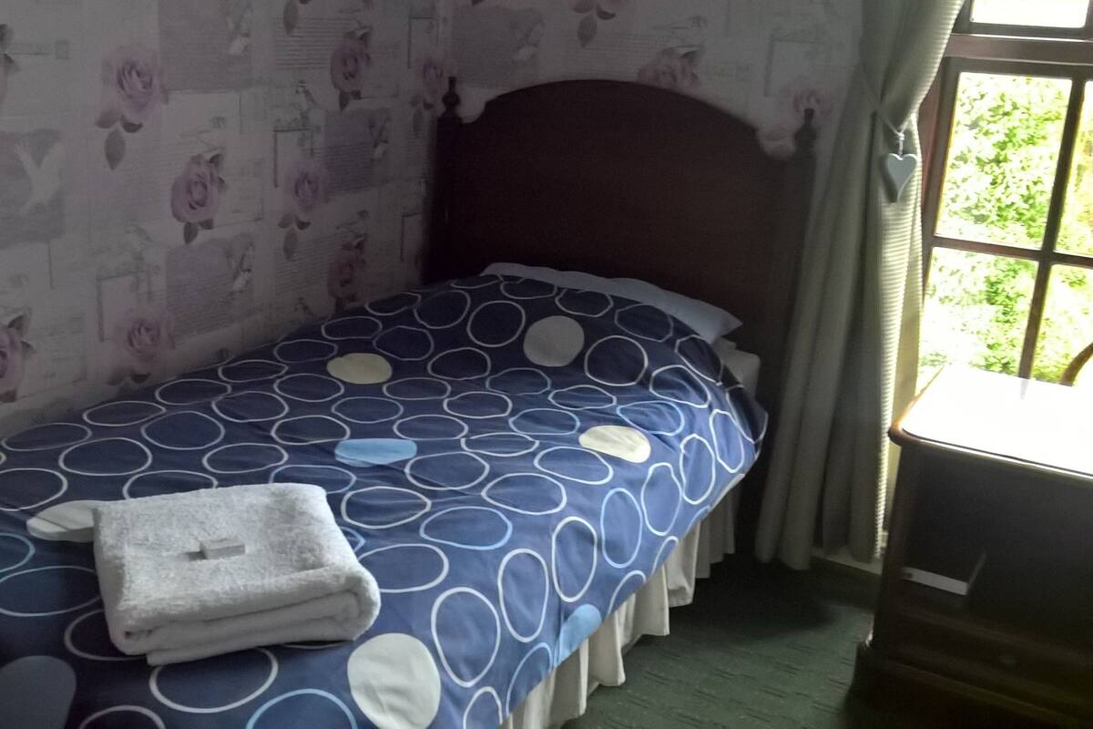 Une petite chambre avec un lit simple, recouverte d’une courtepointe à pois bleus et blancs, un oreiller blanc et une serviette blanche pliée posée sur le lit. Les murs sont recouverts d’un papier peint à motifs floraux et une fenêtre offre une vue sur la verdure à l’extérieur.