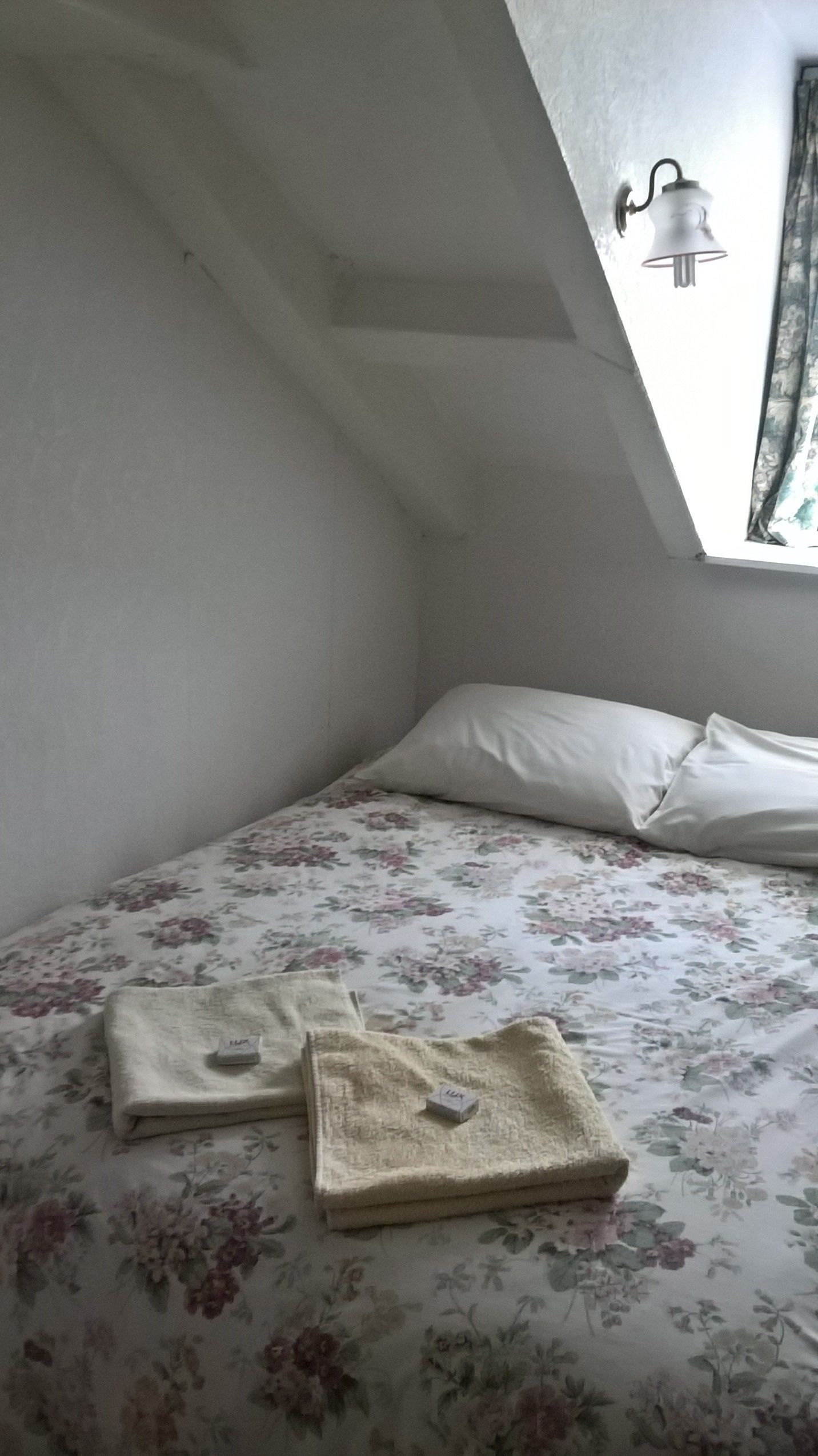 Une petite pièce aux murs légèrement inclinés, avec un lit simple, un oreiller et une table de chevet sur laquelle se trouve une lampe.